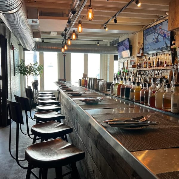 Interior of Wicked Sisters bar with stools, bottles, and TVs showing sports, bathed in natural light.