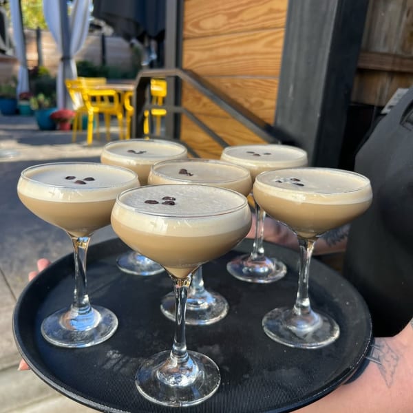 A server carries a tray of six espresso martinis with coffee bean garnishes on a patio.