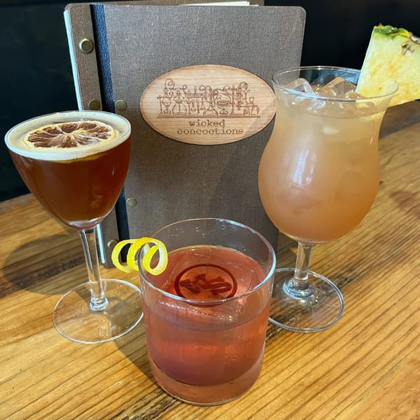 Three Wicked Sisters cocktails, two garnished with citrus, on a wooden table with a menu.