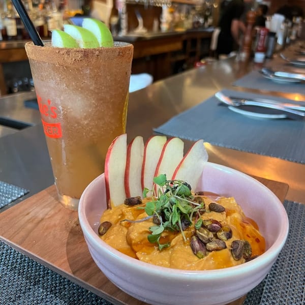 A Wicked Sisters cocktail with apple slices and a bowl of hummus with pistachios and microgreens.