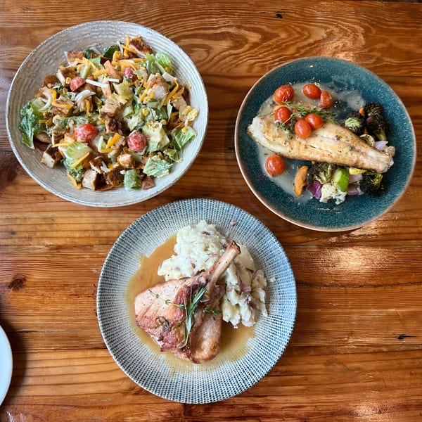 Overhead view of three American dishes: a chicken salad, fish with roasted vegetables, and a pork chop with mashed potatoes.