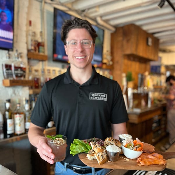 Smiling Wicked Sisters staff member holding a cocktail and a bagel and lox board.