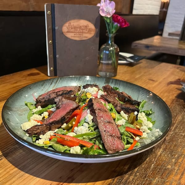 A Wicked Sisters steak salad with greens, peppers, corn, and feta cheese on a wooden table.